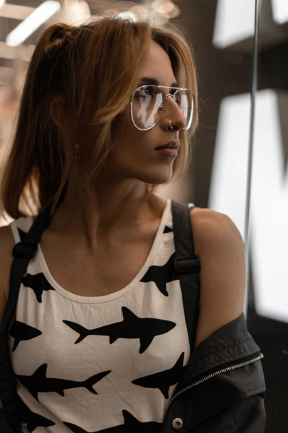Portret van een mooie jonge hipster vrouw in Vintage glazen met een oorbel in de neus met een trendy kapsel in een modieuze t-shirt met een patroon in de buurt van de glazen vitrine in een winkelcentrum. Urban meisje model. - Foto, afbeelding