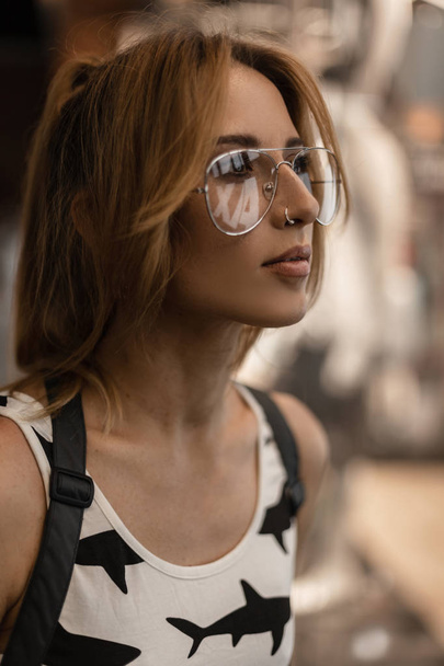 Portrait of a beautiful young hipster woman in trendy glasses with an earring in nose with a stylish hairstyle in a fashionable t-shirt with a pattern near the glass showcase in a store. Pretty girl. - Fotó, kép