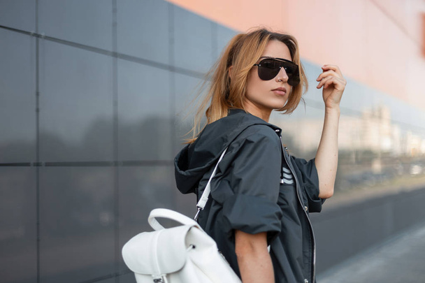 Red-haired pretty young hipster woman in stylish jacket with vintage leather backpack in trendy sunglasses standing near modern building in the city on a summer day. Urban beautiful girl model. - Photo, Image