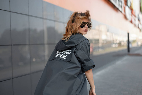 Die schöne junge rothaarige Hipster-Frau mit der trendigen schwarzen Sonnenbrille spaziert in einer stylischen langen Modejacke in der Nähe eines grauen Gebäudes durch die Stadt. modernes attraktives Mädchen geht und dreht sich um. - Foto, Bild