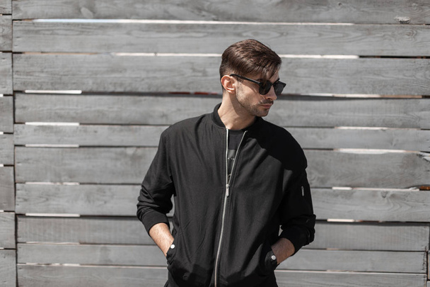 Young stylish hipster man in a vintage jacket in fashionable sunglasses rest on the street on a sunny summer day. Modern handsome guy stands near a gray wooden wall outdoors. Trendy menswear. - Photo, Image