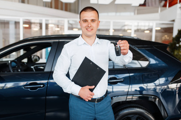 Ο πωλητής δίνει το κλειδί για το νέο αυτοκίνητο στην έκθεση. Αρσενικό πελάτη αγοράζοντας όχημα σε αντιπροσωπεία, πώληση αυτοκινήτων, αυτόματη αγορά - Φωτογραφία, εικόνα