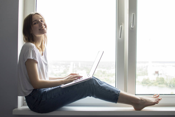 ラップトップで窓の近くに座っている若い女の子は、彼女は窓ガラス、女性フリーランス、テキストのためのコピースペースに自宅でコンピュータを使用しています - 写真・画像