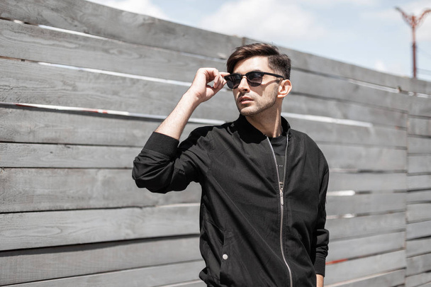 Stylish young hipster man in a vintage black jacket in fashionable sunglasses with a trendy hairstyle relaxes near a wooden fence on a bright sunny day.American urban guy enjoys the sunshine outdoors. - Photo, Image