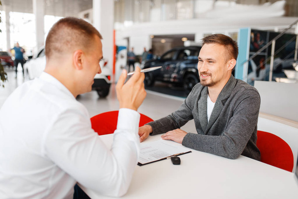 Manager and buyer make out the purchase of new car in showroom. Male customer buying vehicle in dealership, automobile sale - Photo, image