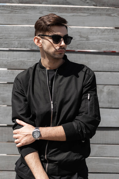 Handsome young hipster man with fashionable hairstyle in trendy black jacket in a stylish dark sunglasses poses near a gray vintage wall in a sunny summer day. Attractive brunette guy outdoors. - Photo, Image