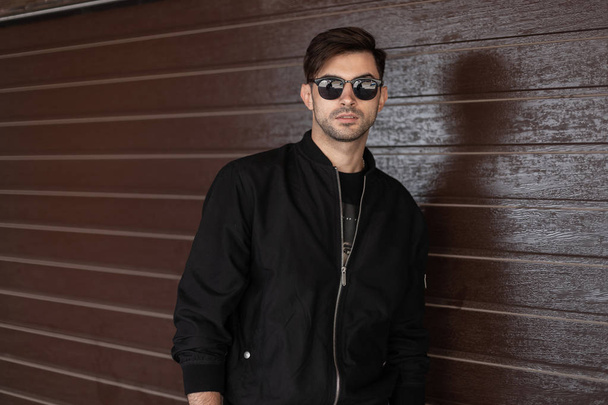 Young attractive hipster man in stylish black jacket in trendy sunglasses with fashionable hairstyle relaxes near brown vintage wall on the street. Modern handsome guy model. Men's clothing. Style. - Photo, image