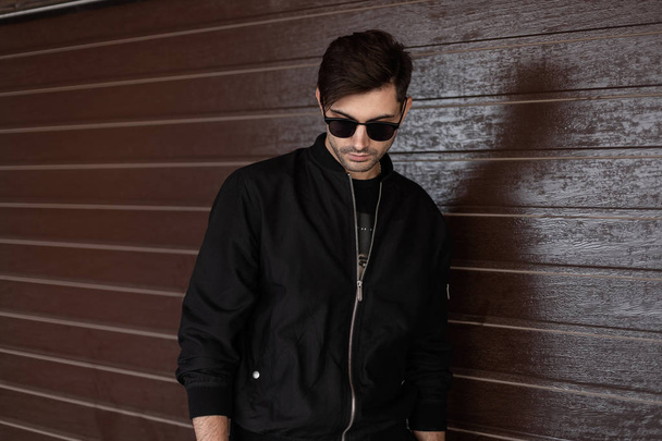Jonge modieuze hipster man in stijlvolle zonnebrillen in een modieuze zwarte jas met een trendy kapsel is rusten in de buurt van een bruine muur van planken in de stad. Aantrekkelijke brunette man. - Foto, afbeelding