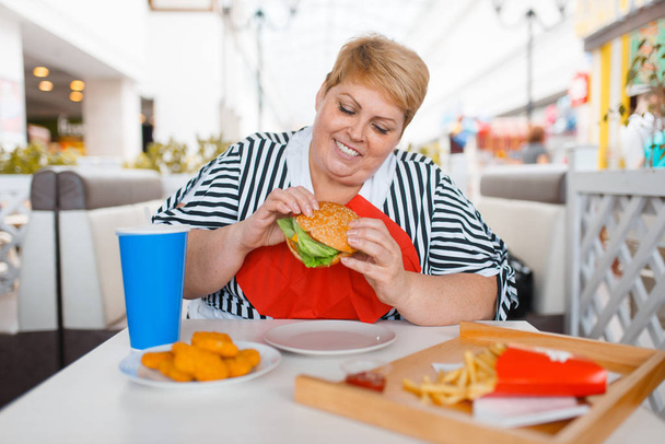 Tlustá žena jedla jídlo v nákupním středisku. Nadváha ženské osoby u stolu s nevyžádaným obědem - Fotografie, Obrázek