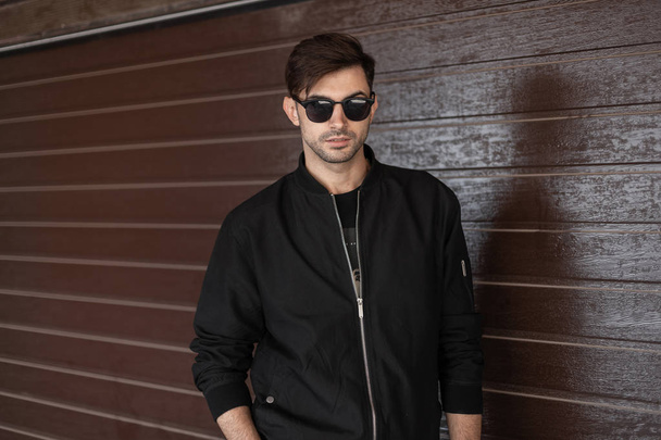 Handsome nice young hipster man in a fashionable black jacket in stylish sunglasses is posing outdoors near a brown wooden vintage wall. Urban guy resting on the street. Street style. Trendy menswear. - Fotografie, Obrázek