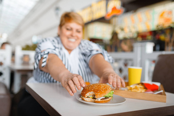 Tlustá žena pojímá vysoké kalorické jídlo v nákupní restauraci. Nadváha ženské osoby u stolu s nevyžádanou večeří, problém s obezitou - Fotografie, Obrázek
