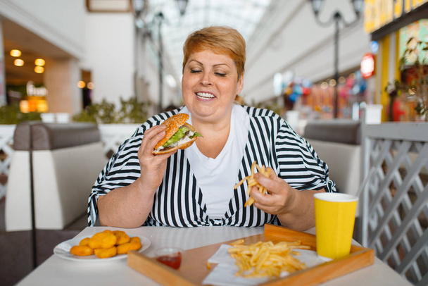 Толстая женщина ест высокую калорийную пищу в ресторане быстрого питания. Избыточный вес женщины за столом с нездоровым ужином, проблемы ожирения
 - Фото, изображение