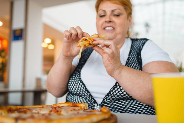 Mujer gorda comiendo pizza con papas fritas en el restaurante del centro comercial, comida poco saludable. Persona femenina con sobrepeso en la mesa con cena basura, problema de obesidad
 - Foto, imagen