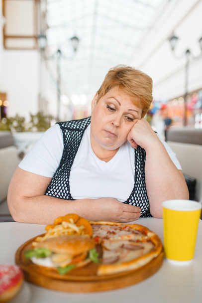 Tučná žena pojímá pizzu v restauraci s občerstvovací restaurací, nezdravé jídlo. Nadváha ženské osoby u stolu s nevyžádanou večeří, problém s obezitou - Fotografie, Obrázek