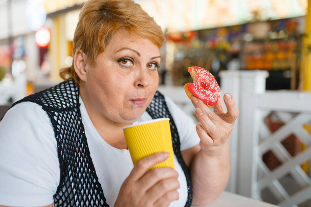 Tlustá žena pojímá koblihy v restauraci s občerstvovací restaurací, nezdravé jídlo. Nadváha ženské osoby u stolu s nevyžádanou večeří, problém s obezitou - Fotografie, Obrázek