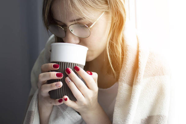 jeune fille couverte d'une couverture, tient une tasse dans ses mains et boit du thé, elle tombe malade et est traitée à la maison, gros plan
 - Photo, image