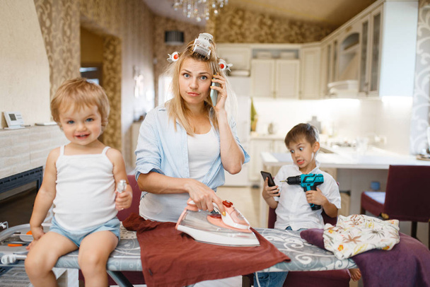 Housewife talking by phone, kids fooling around on the kitchen. Woman with children playing at home together. Female person with daughter and son in their house - Foto, Bild
