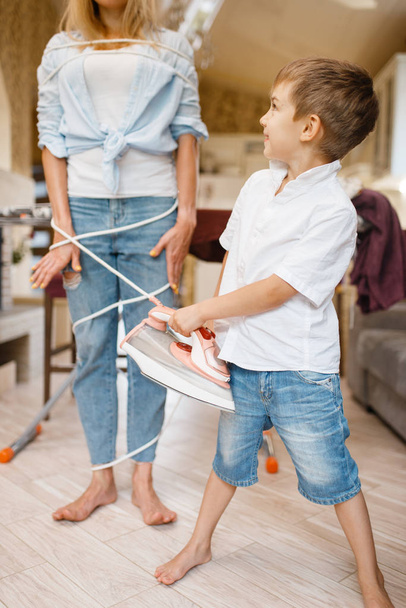 A little boy wrapped a housewife with wires. Woman doing housework at home. Female person with her playful son fooling around in the house - Photo, Image