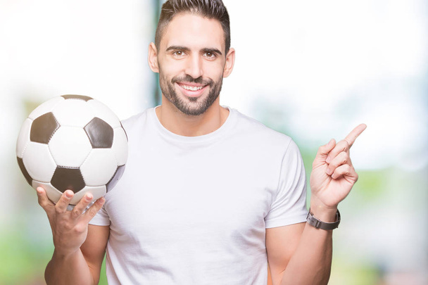 Junger Mann hält Fußballball über isoliertem Hintergrund und zeigt glücklich mit Hand und Finger zur Seite - Foto, Bild