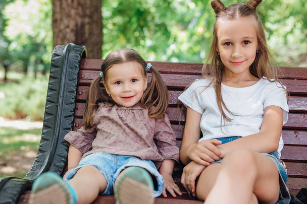 Two cute little girls smiling and sitting on the wooden bench in park - Foto, Imagem