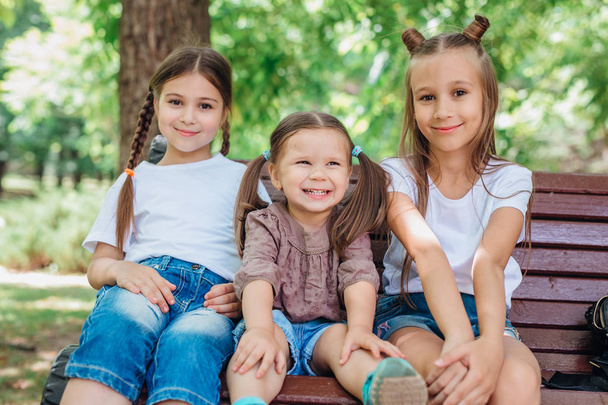 Τρία χαριτωμένα κοριτσάκια χαμογελούν και κάθονται στο ξύλινο παγκάκι στο καλοκαιρινό πάρκο - Φωτογραφία, εικόνα