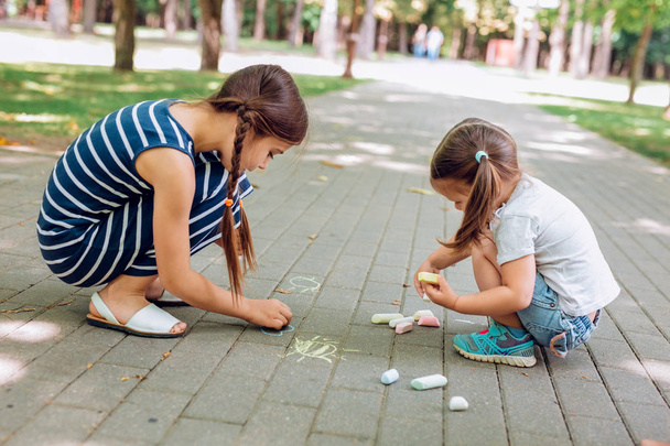Dwie śliczne dziewczynki siedzą i rysują kredą na asfalcie w parku. - Zdjęcie, obraz