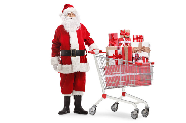 Ganzes Porträt von Weihnachtsmann, der mit Geschenken in einem Einkaufswagen auf weißem Hintergrund steht - Foto, Bild