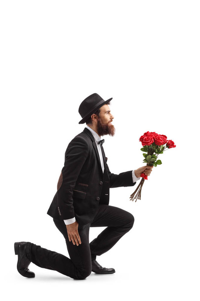 Full length profile shot of a bearded man kneeling and holding red roses isolated on white background - 写真・画像