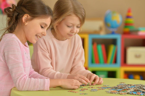 Cute little girls collecting puzzle pieces while sitting at table - Foto, Imagen