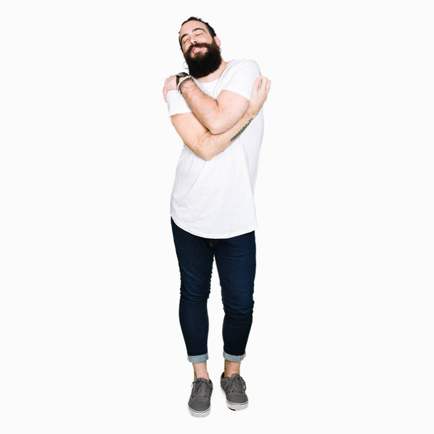 Young hipster man with long hair and beard wearing casual white t-shirt Hugging oneself happy and positive, smiling confident. Self love and self care - Photo, Image