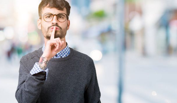 Νέος όμορφος έξυπνος άντρας φορώντας γυαλιά σε απομονωμένο φόντο ζητώντας να είναι ήσυχος με το δάχτυλο στα χείλη. Σιωπή και μυστική αντίληψη. - Φωτογραφία, εικόνα