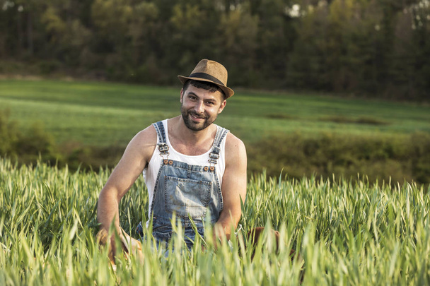 Bearded smiling young man wearing a hat sitting on field while looking camera - Foto, Imagem