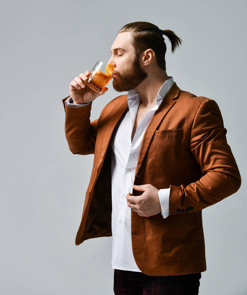 Young stylish bearded man in fashion suit and white shirt tries takes a sip of a delicious cocktail drink with orange - Foto, Imagen