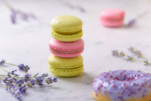 Three macarons and sprigs lavender in the center of the image, one lilac donut in front, one pink macaron behind, on white marble table - Foto, Imagem