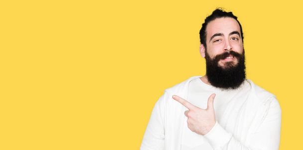 Young man with long hair and beard wearing sporty sweatshirt cheerful with a smile of face pointing with hand and finger up to the side with happy and natural expression on face - Foto, immagini
