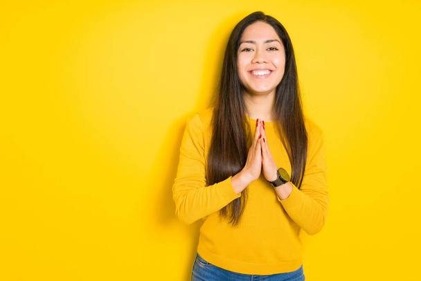 Красива брюнетка над жовтим ізольованим фоном молиться руками, просячи прощення посміхаючись впевнено
. - Фото, зображення
