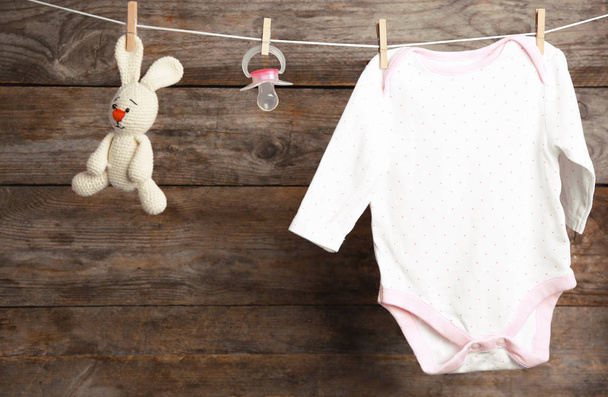 Bodysuit, pacifier and toy bunny on laundry line against wooden background. Baby accessories - Zdjęcie, obraz