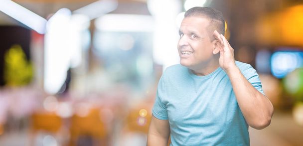 Arabischer Mann mittleren Alters in blauem T-Shirt vor isoliertem Hintergrund, der lächelnd mit der Hand über dem Ohr Gerüchten oder Klatsch lauscht. Konzept für Gehörlosigkeit. - Foto, Bild