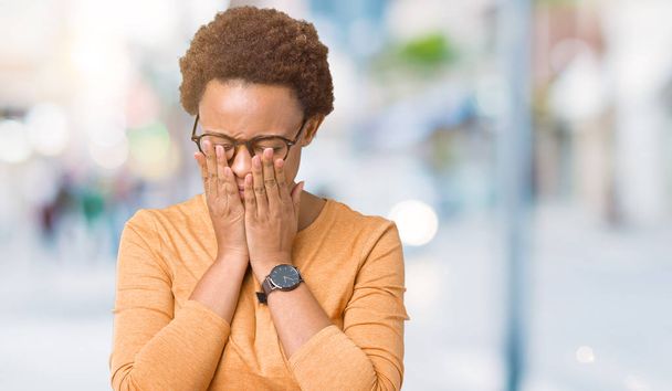 Молодая красивая африканская американка в очках на изолированном фоне с грустным лицом, прикрывающим лицо руками во время плача. Концепция депрессии
. - Фото, изображение