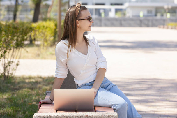 Beautiful woman sitting on a park bench using a laptop. - Фото, зображення