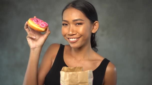 Portrét nádherné mladé usmívající se ženy v černém tričku jíst koblihu nad betonovým pozadím - Záběry, video
