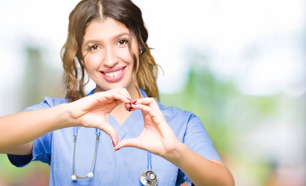 Mladá ošetřující žena, která nosí na lékařské uniformě, se usmívá láskou a rukou se mu zobrazuje symbol srdce a tvar. Romantický koncept. - Fotografie, Obrázek