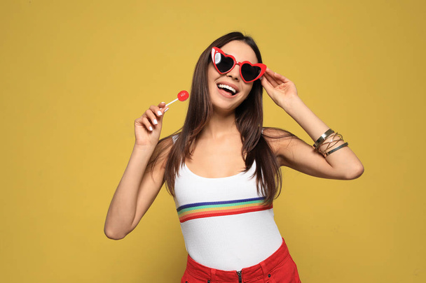 Portrait of beautiful young woman with heart shaped sunglasses and lollipop on color background - Photo, image