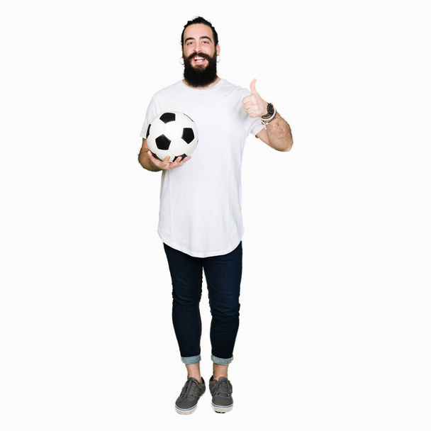 Giovane uomo con i capelli lunghi e orso in possesso di pallone da calcio felice con grande sorriso facendo segno ok, pollice in su con le dita, segno eccellente
 - Foto, immagini