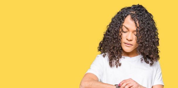 Young beautiful woman with curly hair wearing white t-shirt Checking the time on wrist watch, relaxed and confident - Photo, Image