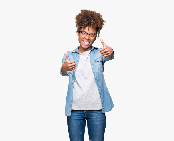 Piękne młode african american Kobieta w okularach na na białym tle zatwierdzające robi pozytywny gest ręką, kciuki, uśmiechając się i szczęśliwy dla sukcesu. Patrząc na kamery, zwycięzca gest. - Zdjęcie, obraz
