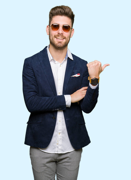 Jonge knappe elegante man dragen zonnebril en mode blazer glimlachend met blij gezicht kijken en naar de kant met duim omhoog. - Foto, afbeelding