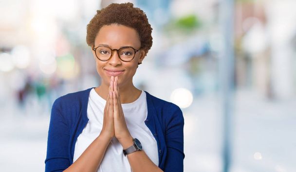 junge schöne afrikanisch-amerikanische Frau mit Brille über isoliertem Hintergrund betet mit den Händen zusammen und bittet um Vergebung, lächelt selbstbewusst. - Foto, Bild