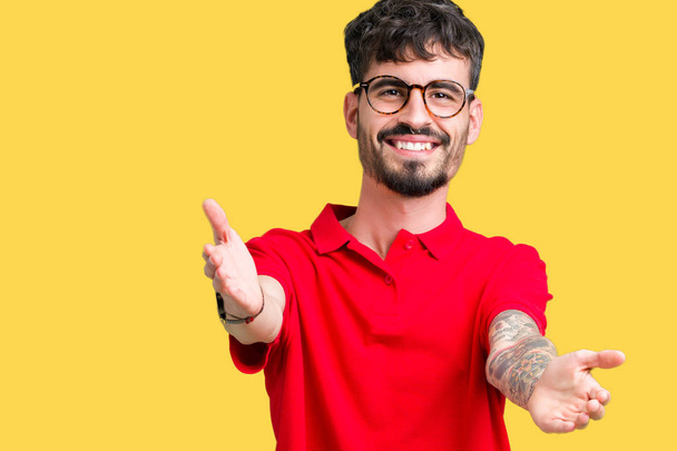 Young handsome man wearing glasses over isolated background looking at the camera smiling with open arms for hug. Cheerful expression embracing happiness. - Photo, Image