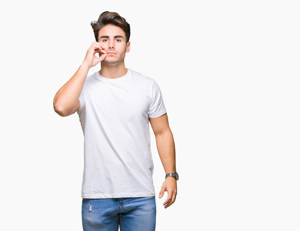 Jonge knappe man dragen witte t-shirt over geïsoleerd achtergrond mond en lippen sluiten als zip met vingers. Geheime en stil, taboe praten - Foto, afbeelding